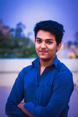 Young man standing outdoors with folded arms, wearing a blue shirt and smiling confidently in front of a blurred background.